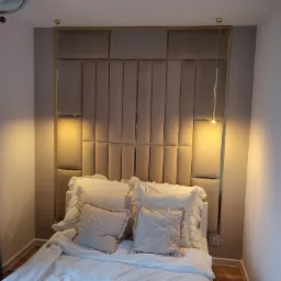 Nowoczesna sypialnia z tapicerowanym panelem ściennym w odcieniach beżu, oświetlonym złotymi lampami wiszącymi i ozdobiona dekoracyjnym żyrandolem.