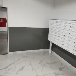 Wnętrze korytarza z białymi płytkami imitującymi marmur na podłodze, szarą lamperią i rzędem białych skrzynek pocztowych z numerami