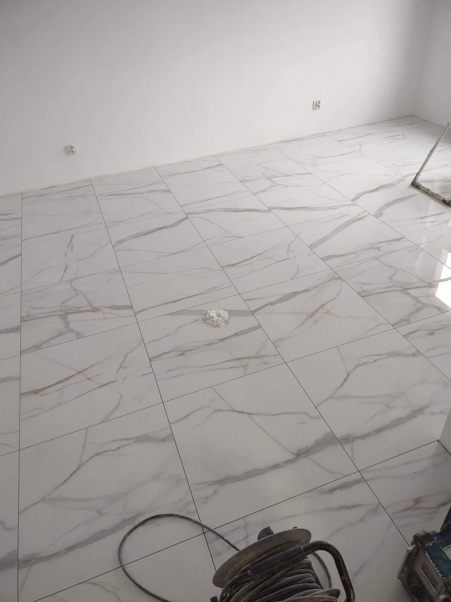 Jasne, marmurowe płytki podłogowe w trakcie układania; widoczny kabel narzędzia oraz resztki zaprawy na powierzchni.