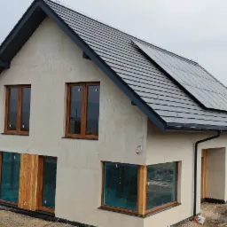 Nowoczesny dom murowany z szarą dachówką i panelami słonecznymi na dachu, surowa elewacja w kolorze kremowym, duże okna z drewnianą stolarką, widok na miasto i góry w tle, teren wokół domu...