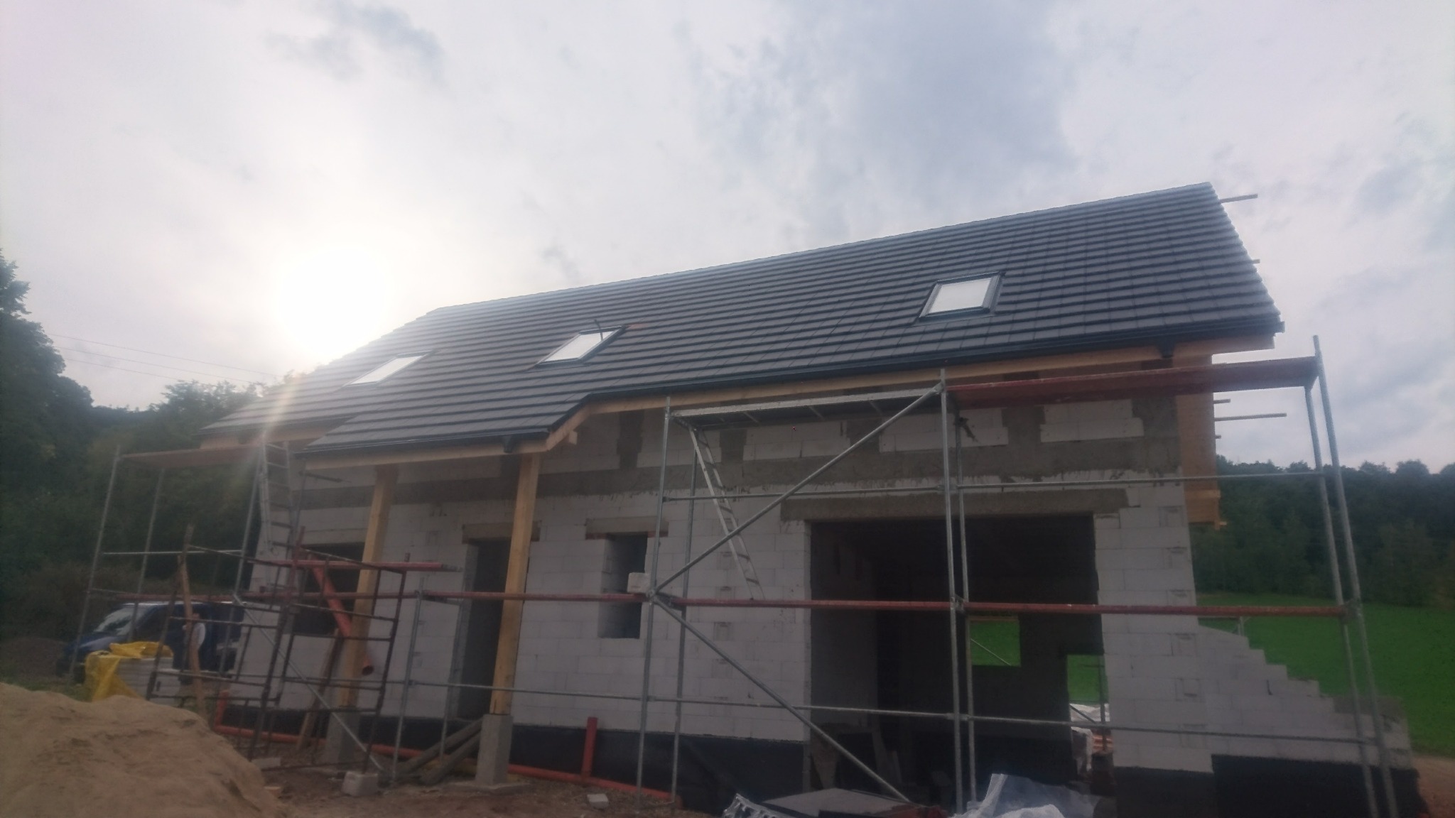 Niedokończony budynek z białego pustaka z czarną hydroizolacją przy gruncie, z częściowo położonym dachem z dachówką i oknami dachowymi, otoczony rusztowaniami i stertą piasku.