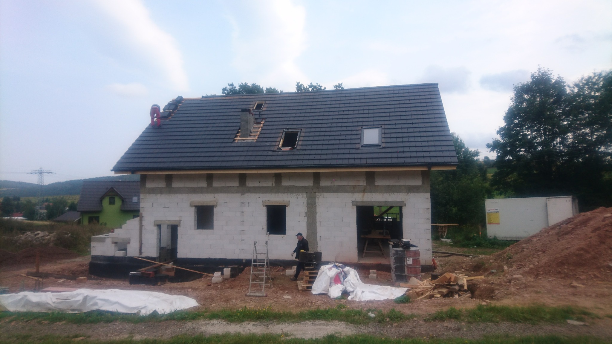 Budowa domu z białych bloczków w Czarnym Borze, widoczni pracownicy na dachu układający ciemną dachówkę, zamontowane okna dachowe, w tle zielony krajobraz.