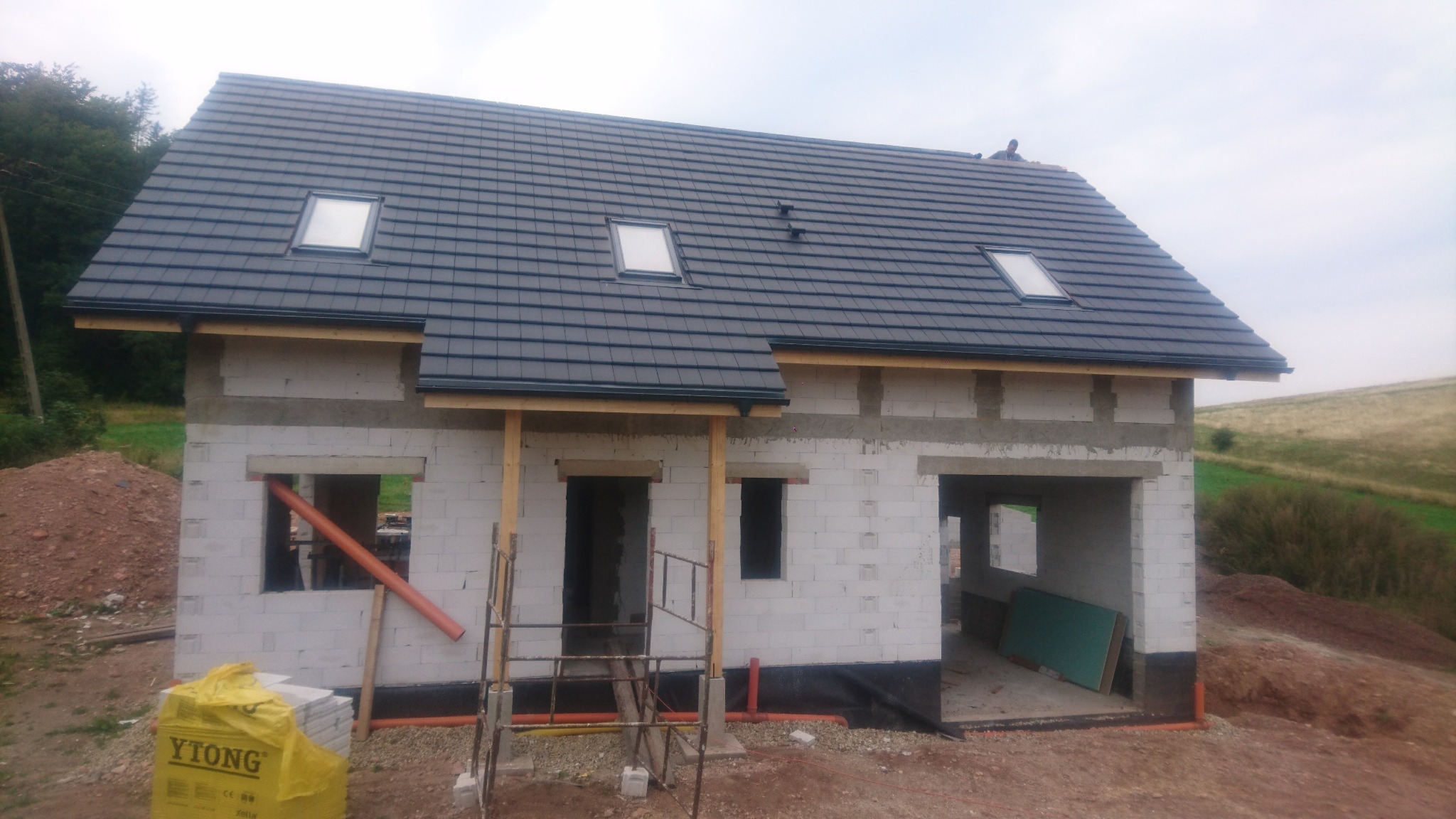 Surowy stan domu jednorodzinnego z białych bloczków Ytong, z zamontowaną szarą dachówką z trzema oknami dachowymi, widoczne rusztowania i materiały budowlane, w tle pagórkowaty krajobraz.