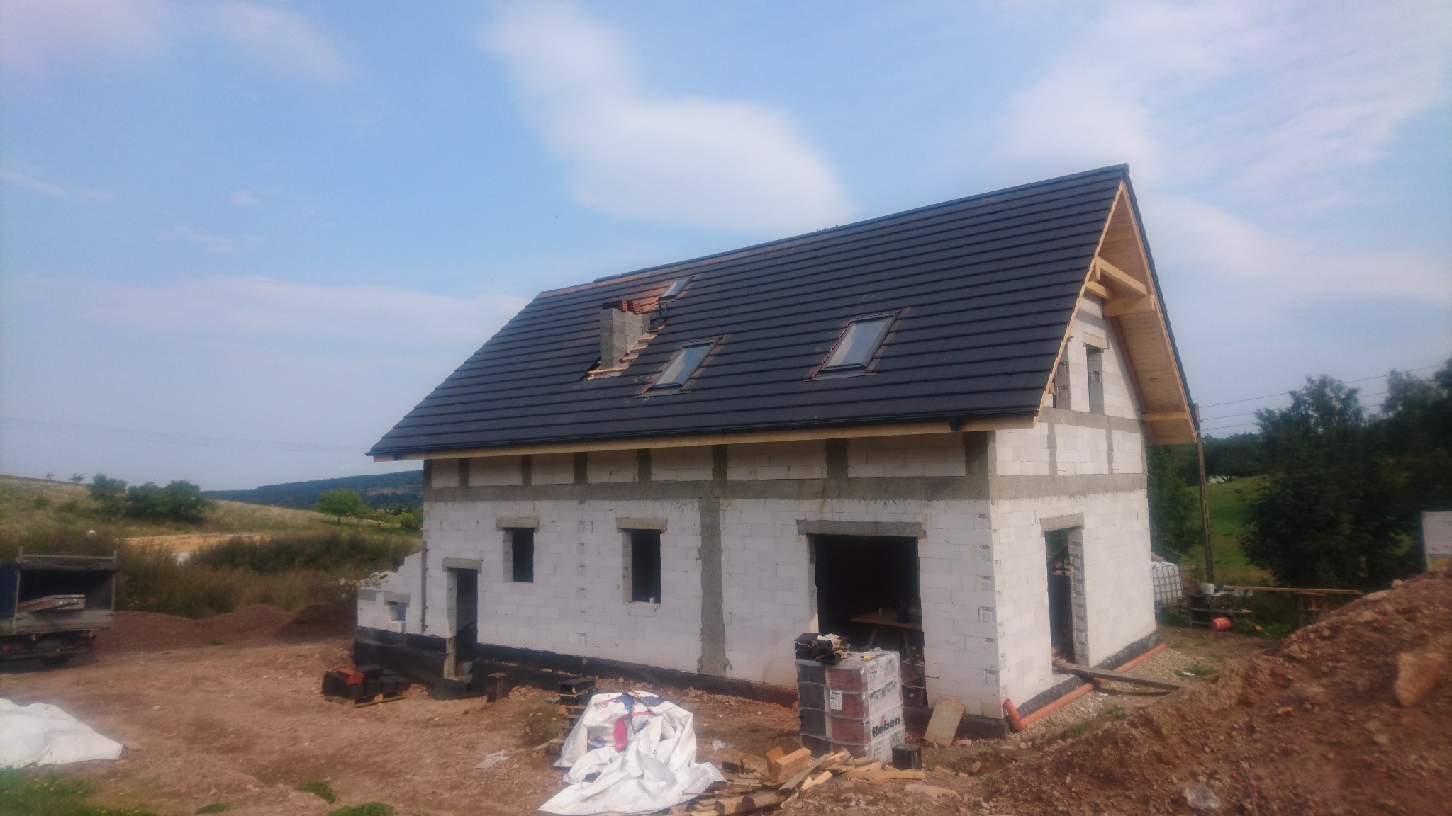 Niedokończony dwukondygnacyjny dom z białych bloczków, z ciemną dachówką, trzema oknami dachowymi i kominem w trakcie budowy, widok z zewnątrz w słoneczny dzień.