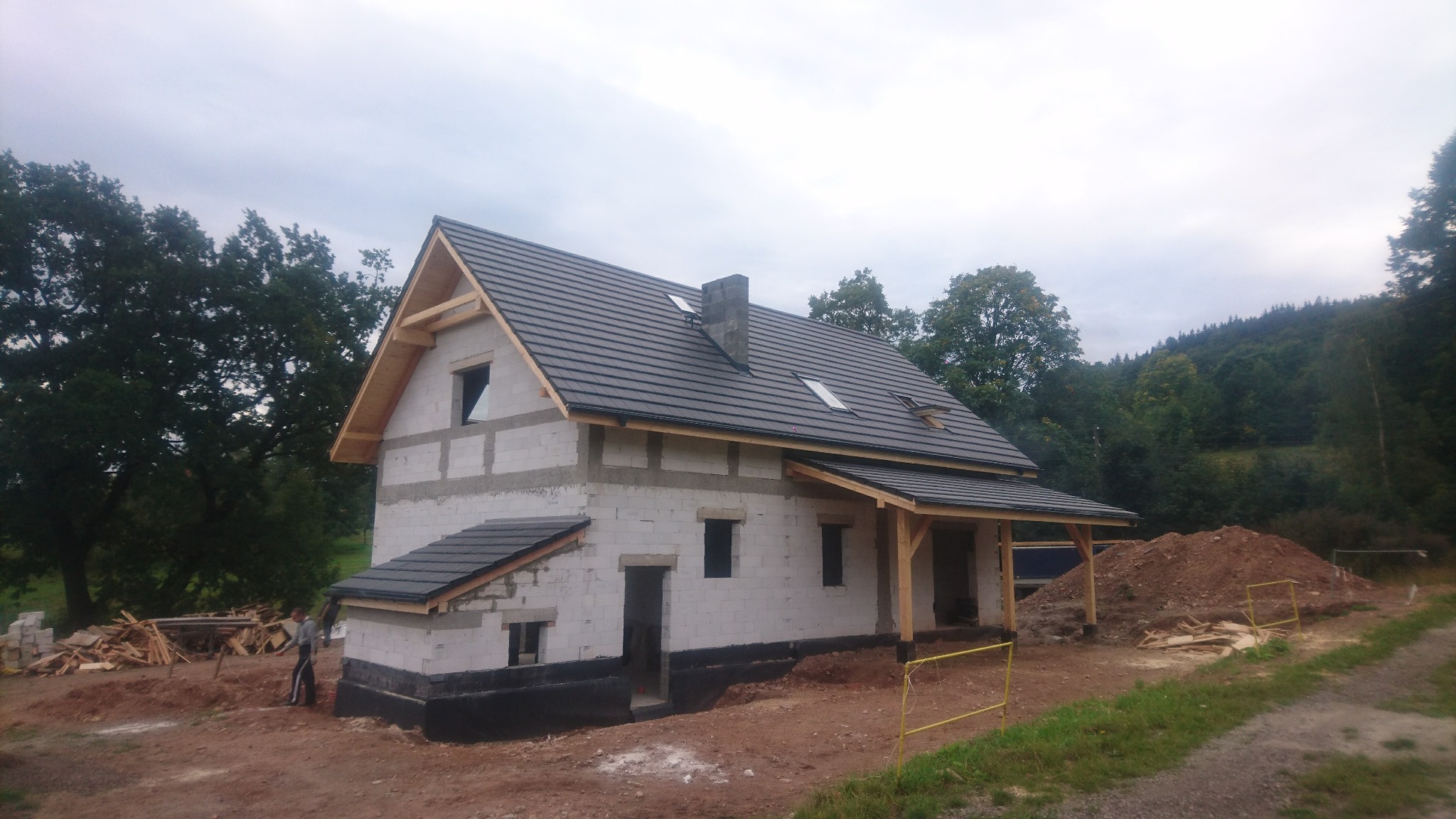 Niedokończony dom z białego bloczka z ciemną dachówką, widoczne drewniane elementy konstrukcyjne dachu i zadaszenia, w tle zalesione wzgórze