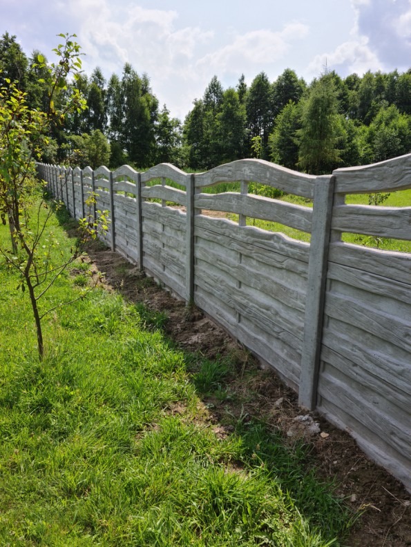 Długi, szary płot z prefabrykatów betonowych imitujących drewno, oddzielający posesję od zieleni drzew w tle, widziany z perspektywy trawnika.