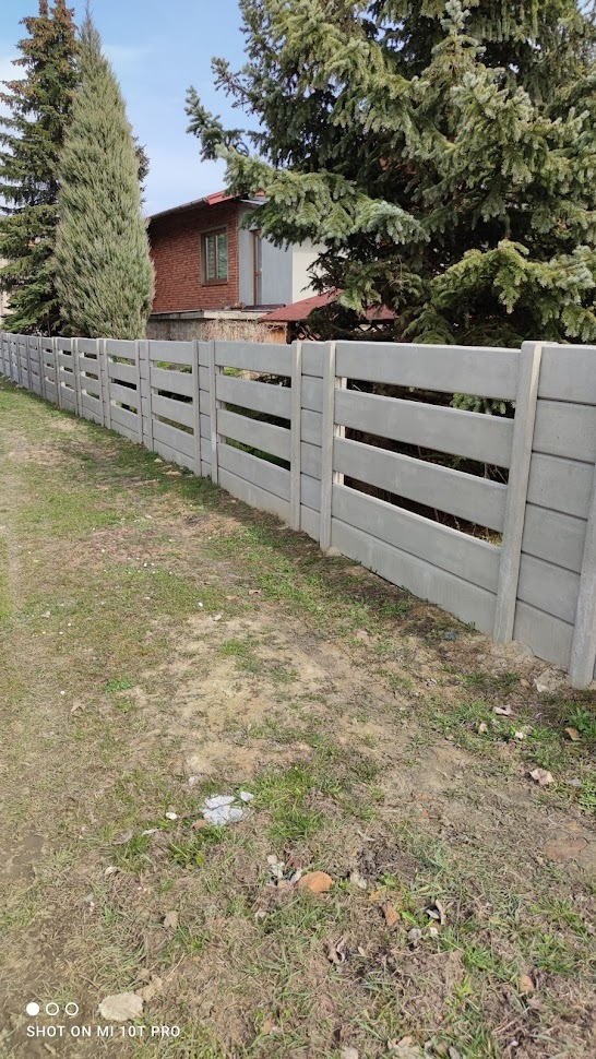 Szary, prefabrykowany płot z betonu z poziomymi szczelinami, oddzielający posesję od drogi gruntowej, w tle dom z czerwonej cegły i wysokie drzewa iglaste.