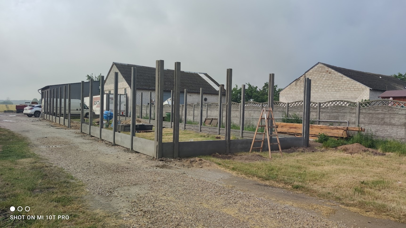 Wznoszenie ogrodzenia betonowego z prefabrykatów: widok na konstrukcję z betonowych słupków i podmurówki, częściowo ukończoną, z drabiną i stosem drewnianych desek w tle, obok istniejącego ogrodzenia.