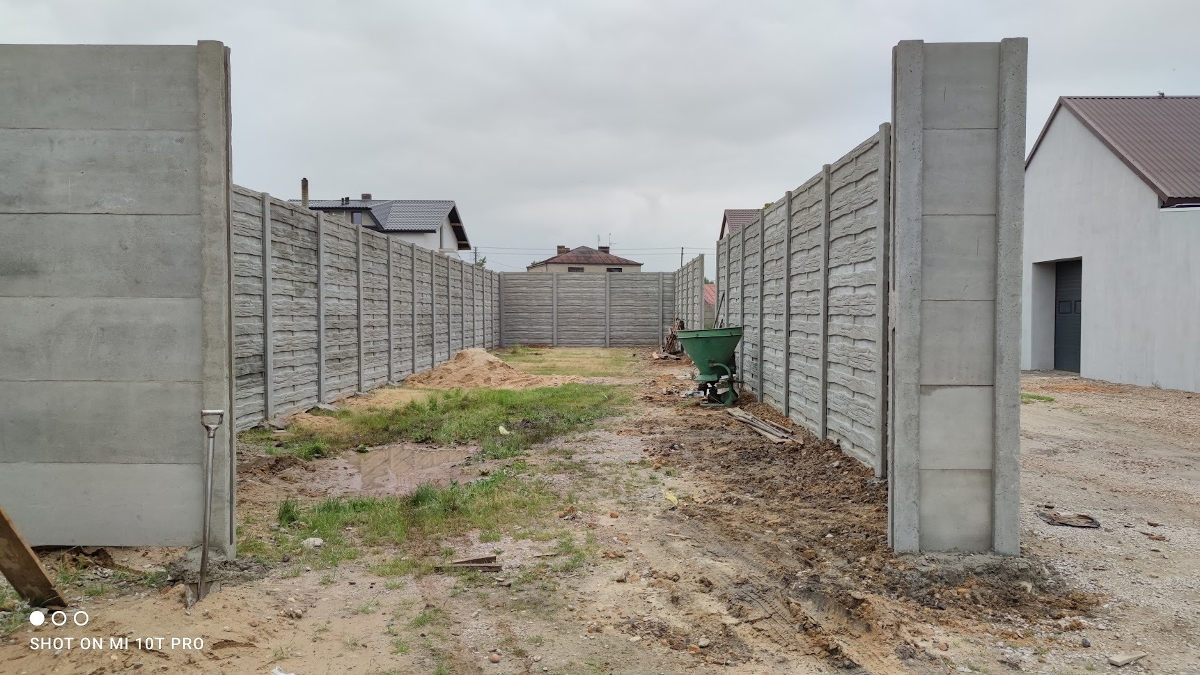 Wnętrze posesji otoczone panelami z imitacją drewna i betonowymi słupkami, widoczny zielony rozsiewacz nawozu i łopata oparta o słupek, w tle domy sąsiadów.