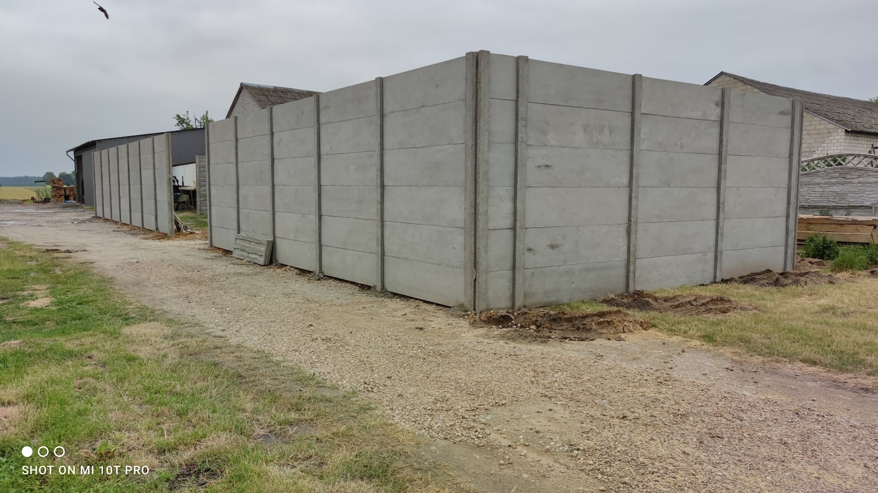 Nowoczesne, modułowe ogrodzenie z prefabrykatów betonowych, widoczne elementy konstrukcyjne i fragment terenu z wykopaną ziemią, sugerujące etap montażu.