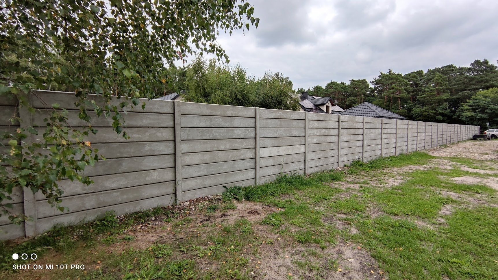 Długi, szary, prefabrykowany płot betonowy z poziomymi panelami, widok z niskiej perspektywy, z fragmentami drzew po lewej stronie i domami w tle.