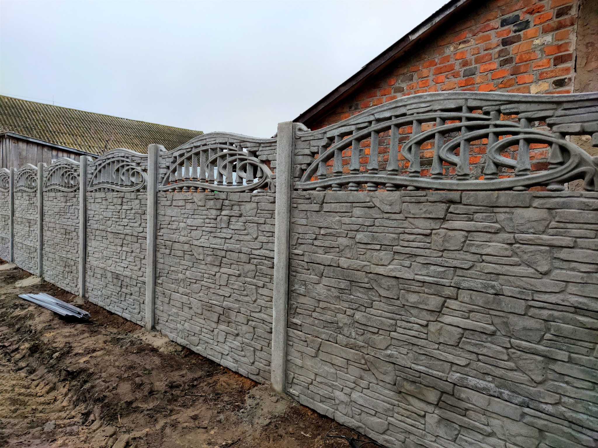 Długi fragment szarego, betonowego ogrodzenia z dekoracyjnym, ażurowym wykończeniem u góry, imitującego kamień, na tle budynku z czerwonej cegły i drewnianego budynku gospodarczego.
