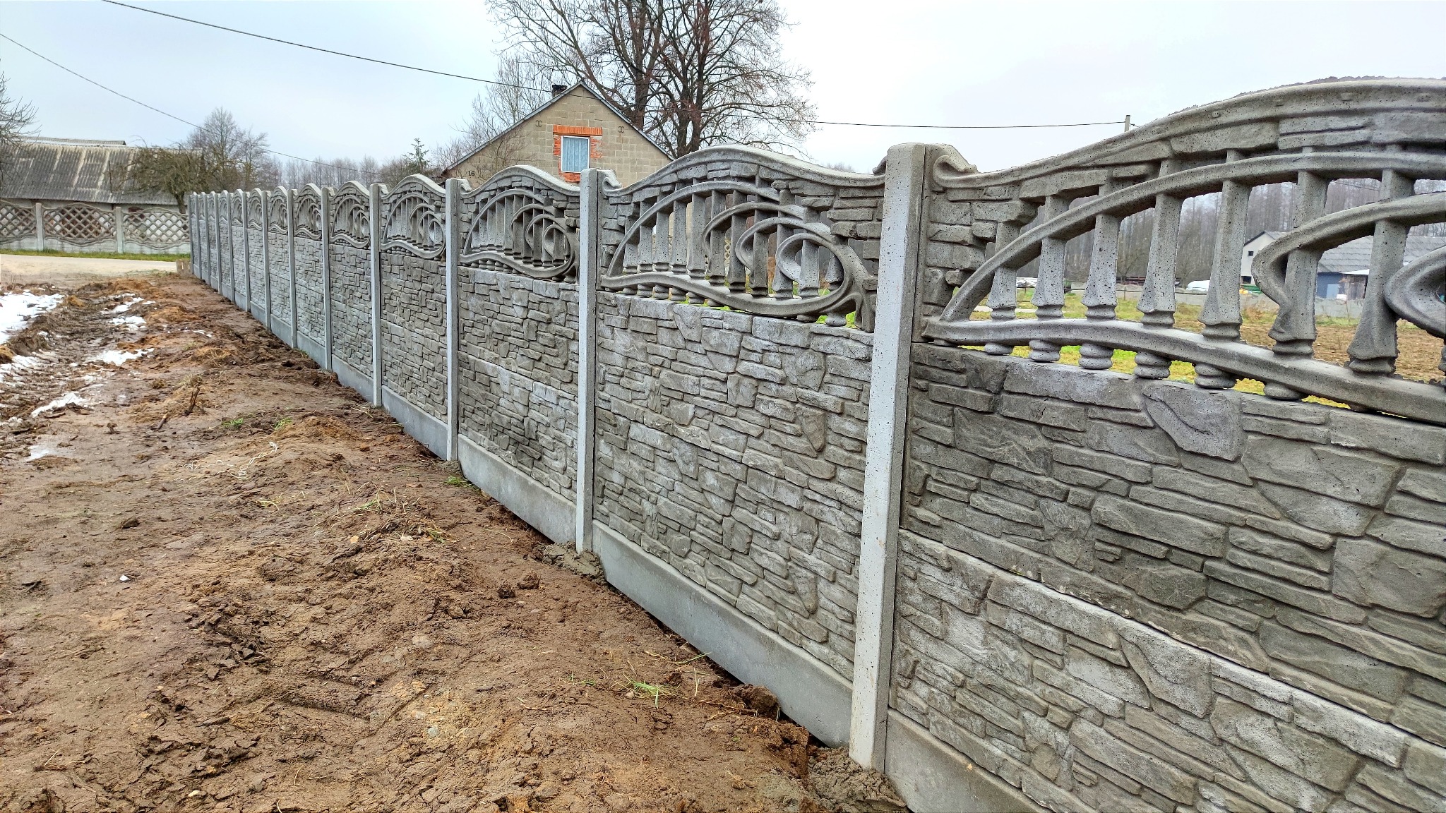 Długi fragment ozdobnego ogrodzenia z prefabrykatów imitujących kamień, widok z poziomu gruntu na błotniste podłoże, domy w tle.