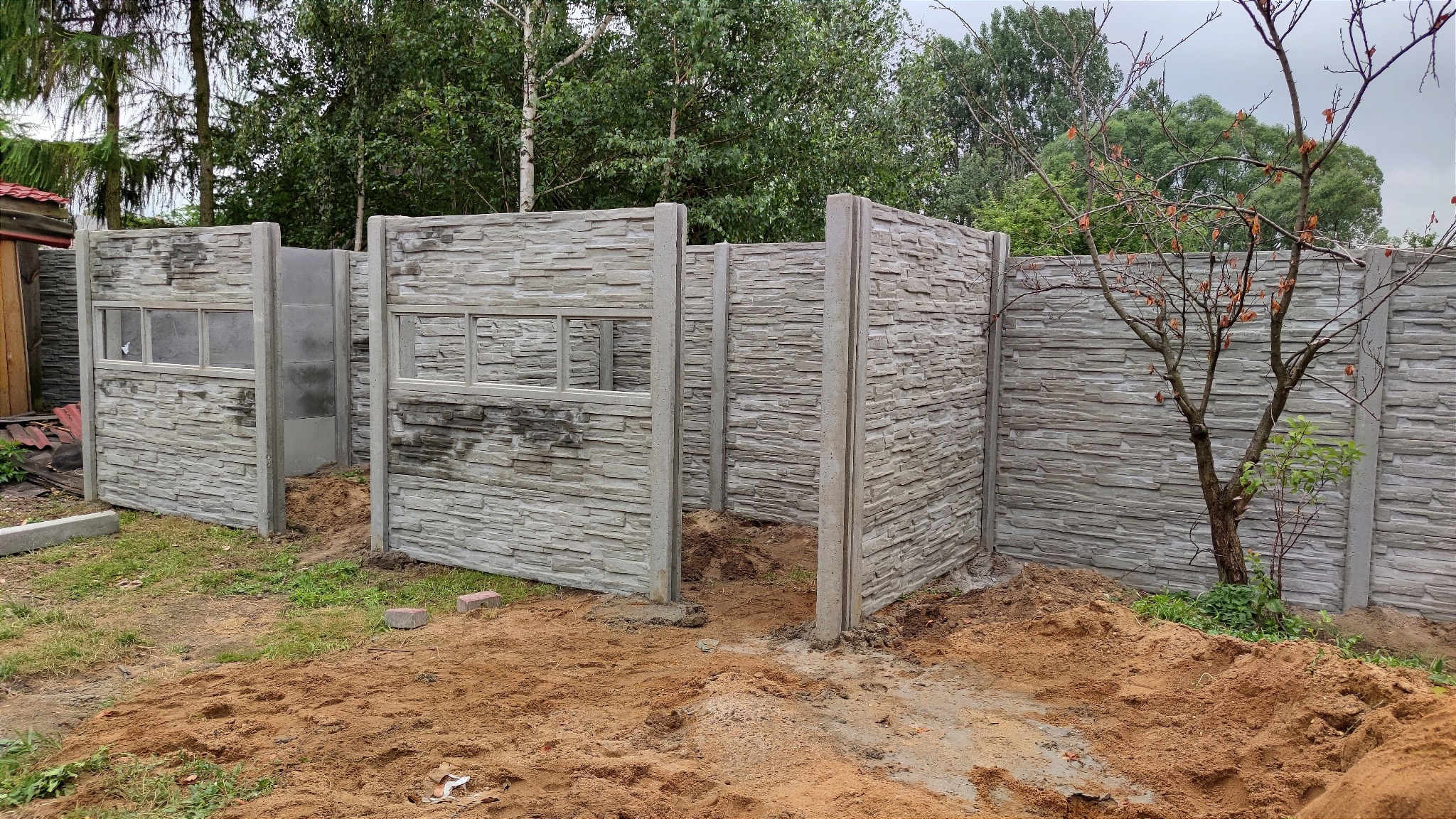 Montaż segmentowego ogrodzenia betonowego imitującego kamień, z otworami okiennymi w panelach, widoczne słupki i panele w trakcie instalacji, rozkopana ziemia w tle.