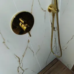 Błyszcząca, złota bateria prysznicowa i słuchawka na ścianie z marmurowymi płytkami w beżowo-białej tonacji, uzupełnione drewnopodobną podłogą w kabinie prysznicowej.