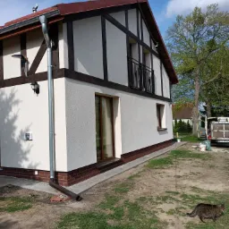 Odnowiona elewacja budynku z elementami drewnianymi, widoczny system rynnowy, kot spacerujący po trawniku i samochód dostawczy w tle.