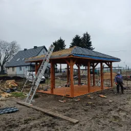 Drewniana altana ogrodowa w trakcie krycia dachu dachówką ceramiczną, widoczna drabina oparta o konstrukcję i pracownik w roboczym ubraniu, w tle dom jednorodzinny i pochmurne niebo.
