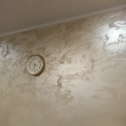 Beżowa ściana wewnętrzna z dekoracyjną fakturą, na której wisi okrągły zegar z jasną tarczą i złotą obudową, wskazujący godzinę 11:53.