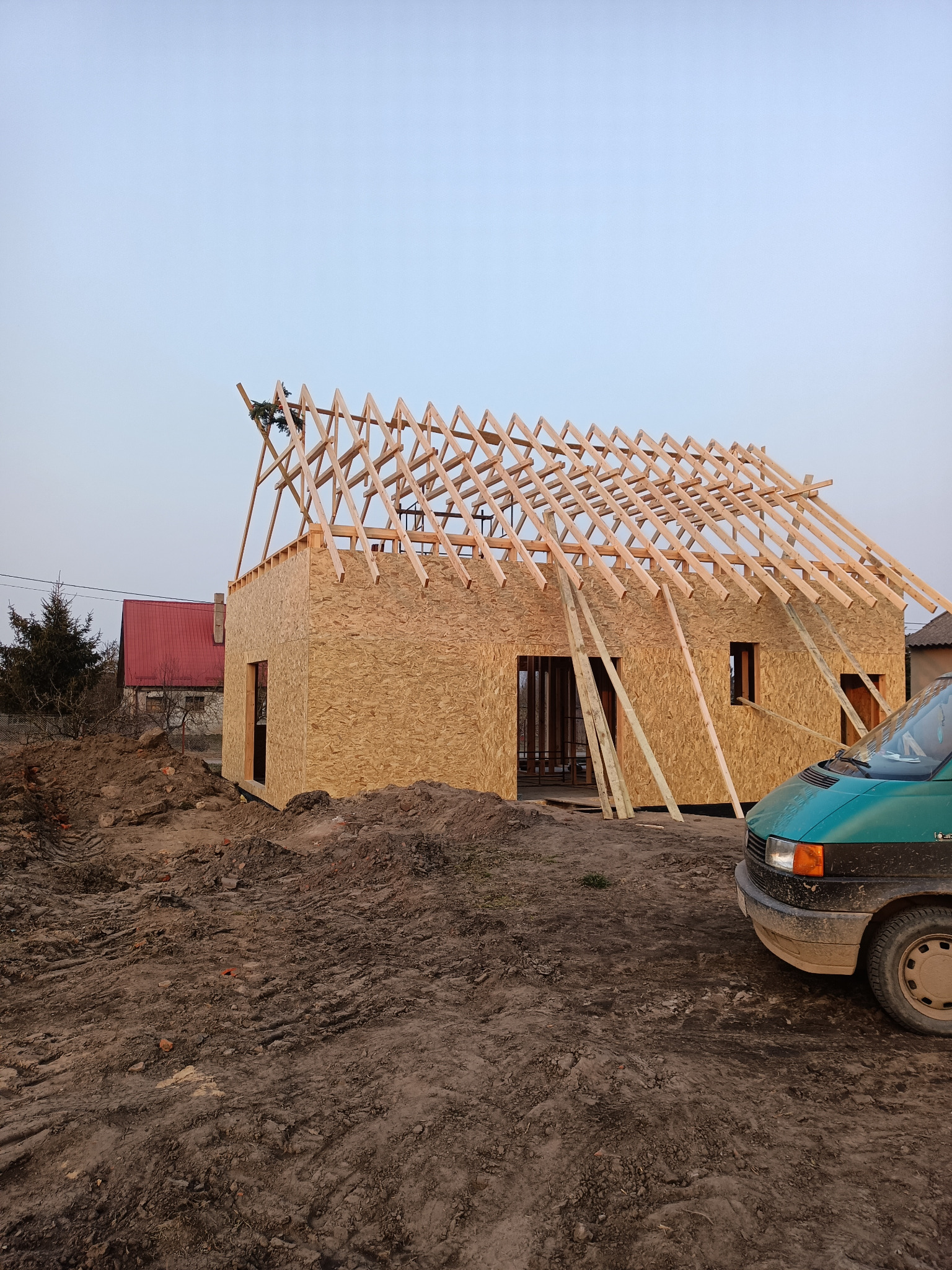 Surowy stan domu szkieletowego z płyt OSB i drewnianą konstrukcją dachu w trakcie budowy, z widocznym zielonym samochodem dostawczym obok.