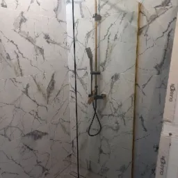 Nowoczesna kabina prysznicowa z marmurowymi płytkami, czarną armaturą i złotymi akcentami na szkle, widoczny odpływ liniowy w podłodze.