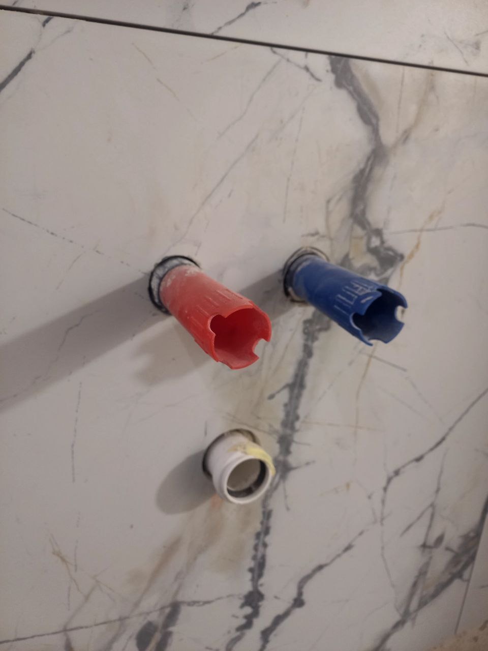 Przygotowane otwory na rury wodne (czerwona i niebieska) oraz kanalizacyjną w płytkach imitujących marmur, żółty klej widoczny w otworze kanalizacyjnym.