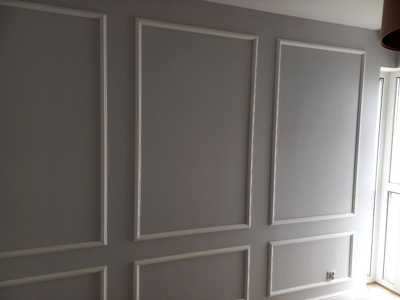 Szara ściana z dekoracyjnymi, białymi panelami w kształcie prostokątów, widoczne okno z jasnym światłem w tle oraz fragment lampy sufitowej.