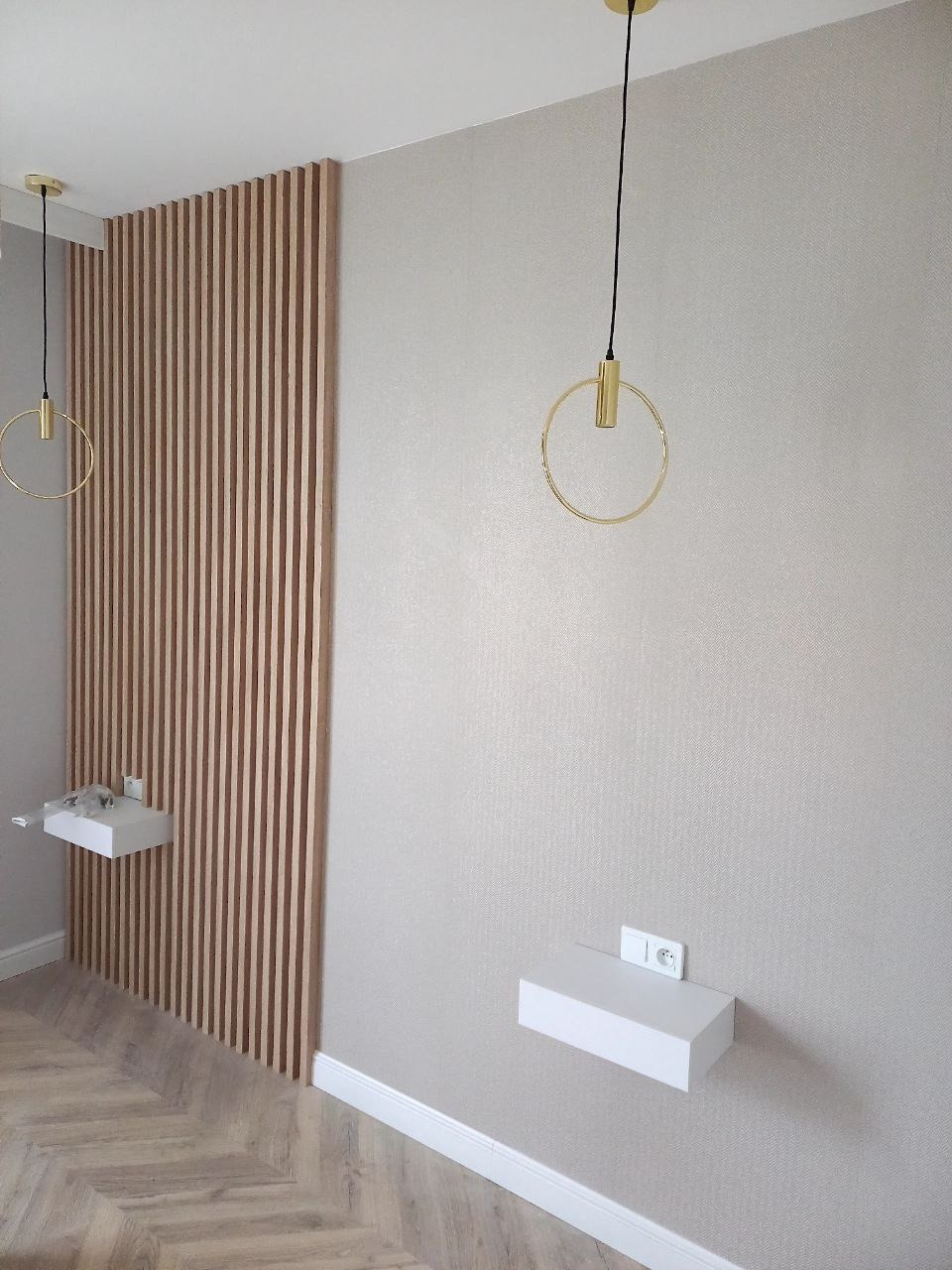 Nowoczesna sypialnia z drewnianą lamelową ścianą, pływającymi białymi szafkami nocnymi z gniazdkami i okrągłymi złotymi lampami wiszącymi.