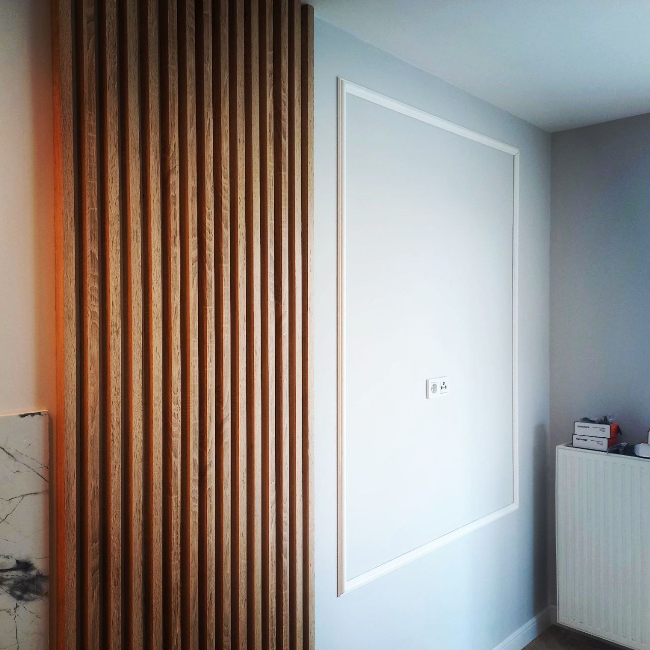 Ściana z drewnianymi lamelami obok białej ściany z dekoracyjną ramą i gniazdkiem elektrycznym, widoczny fragment marmurowego blatu.