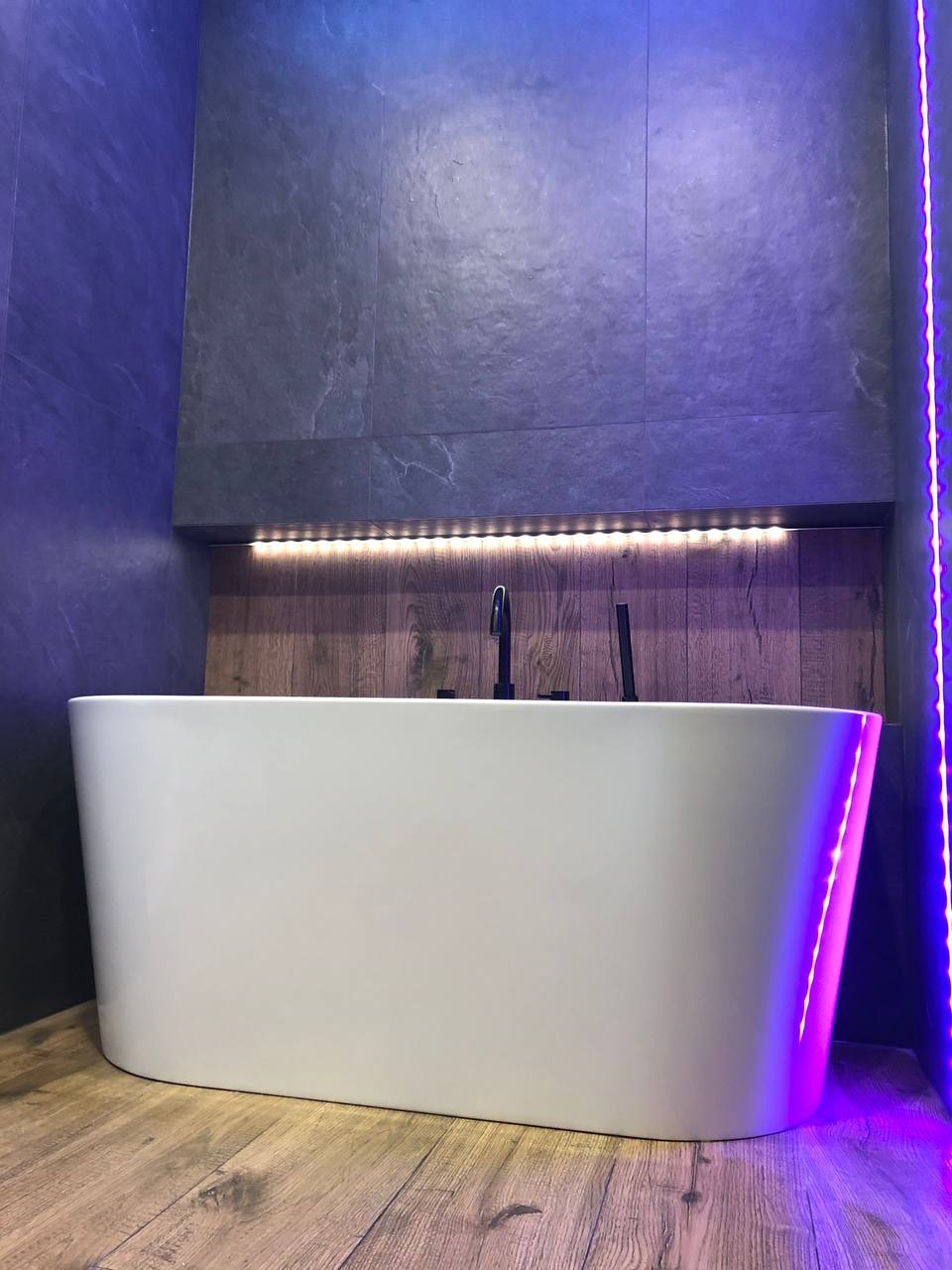 Nowoczesna biała wanna wolnostojąca na drewnopodobnej podłodze, z szarymi płytkami imitującymi kamień na ścianie i akcentami niebieskiego oświetlenia LED.