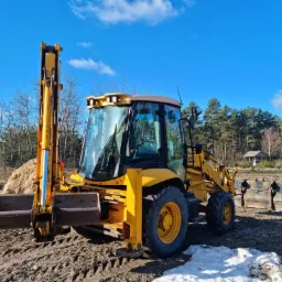 Żółta koparko-ładowarka JCB 3CX na nierównym, częściowo błotnistym terenie z pryzmą piasku w tle, pod błękitnym niebem z chmurami.