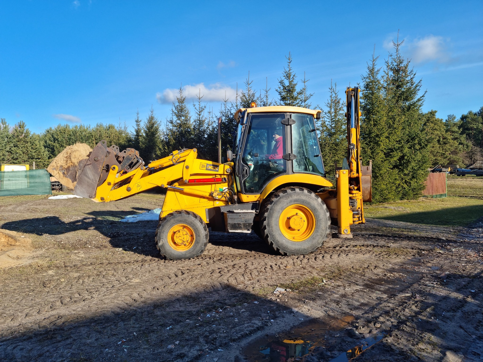 Żółta koparko-ładowarka JCB na placu budowy, z łyżką wypełnioną ziemią, operator w kabinie, w tle drzewa iglaste i błękitne niebo.