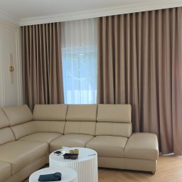 Jakby Luksusowo Studio Dekoracji - Nowoczesny salon z beżową sofą narożną, dwoma białymi stolikami kawowymi z ryflowanymi bokami, drewnianą podłogą w jodełkę i eleganckimi, beżowymi zasłonami oraz białymi firanami.