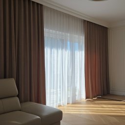Jakby Luksusowo Studio Dekoracji - Plisowane brązowe zasłony i białe firany w nowoczesnym salonie. Słońce rzuca długie cienie na drewnianą podłogę w jodełkę, obok beżowa sofa i designerska lampa.