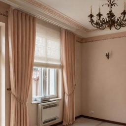 Jakby Luksusowo Studio Dekoracji - Jasnobrzoskwiniowe zasłony z ozdobnymi podpięciami i roleta rzymska w oknie, w pokoju z klasycznym żyrandolem, sztukaterią sufitową i zabezpieczoną podłogą.