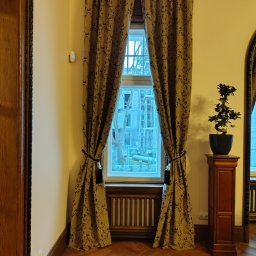 Jakby Luksusowo Studio Dekoracji - Eleganckie, brązowo-złote zasłony z klasycznym wzorem, podpięte ozdobnymi chwostami, w oknie z drewnianym parapetem i parkietem w jodełkę.