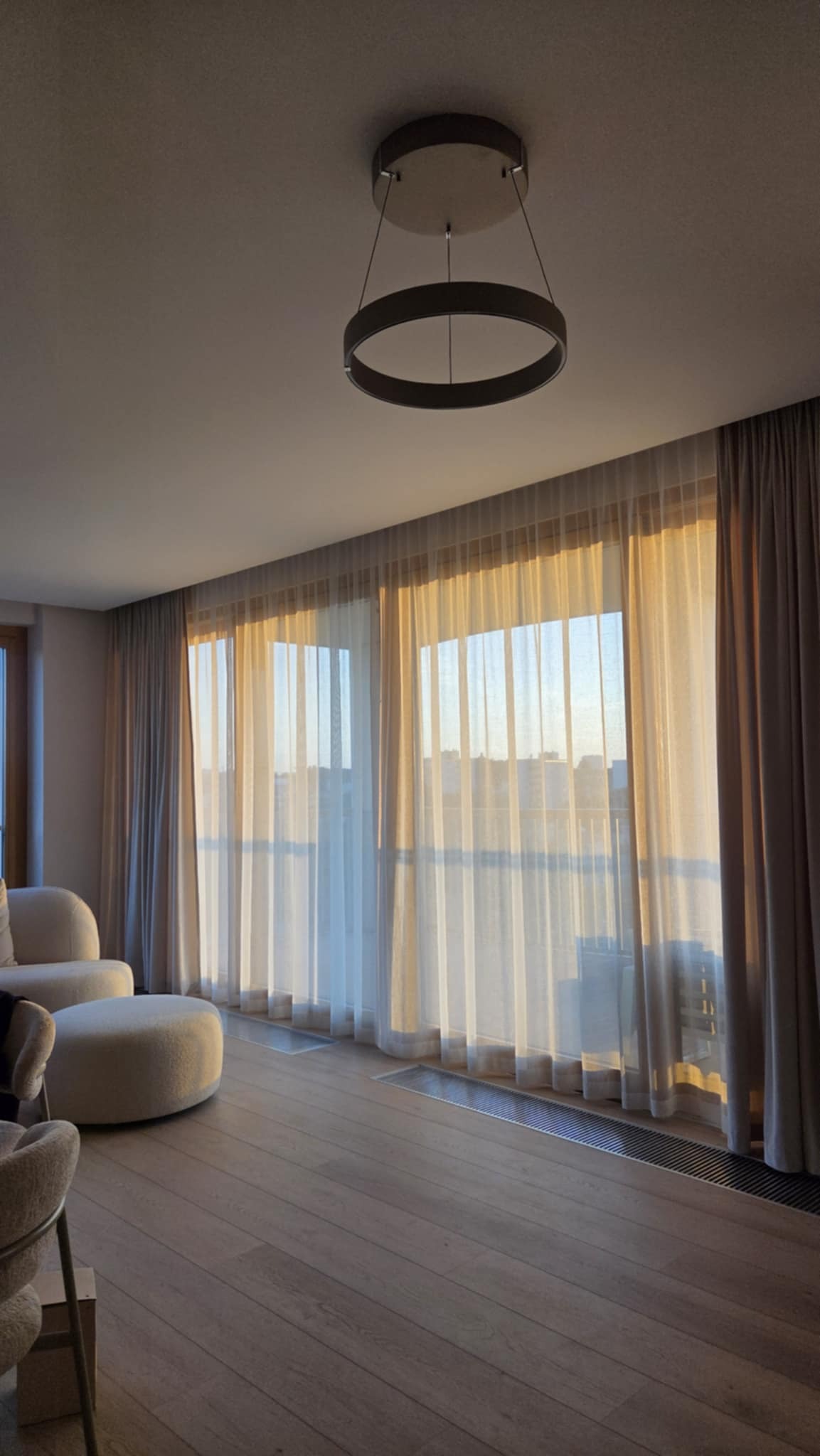 Elegancki salon z beżowymi zasłonami, przez które wpada ciepłe światło słoneczne. Okrągła lampa sufitowa dodaje nowoczesnego akcentu. Widoczne elementy wystroju wnętrza.