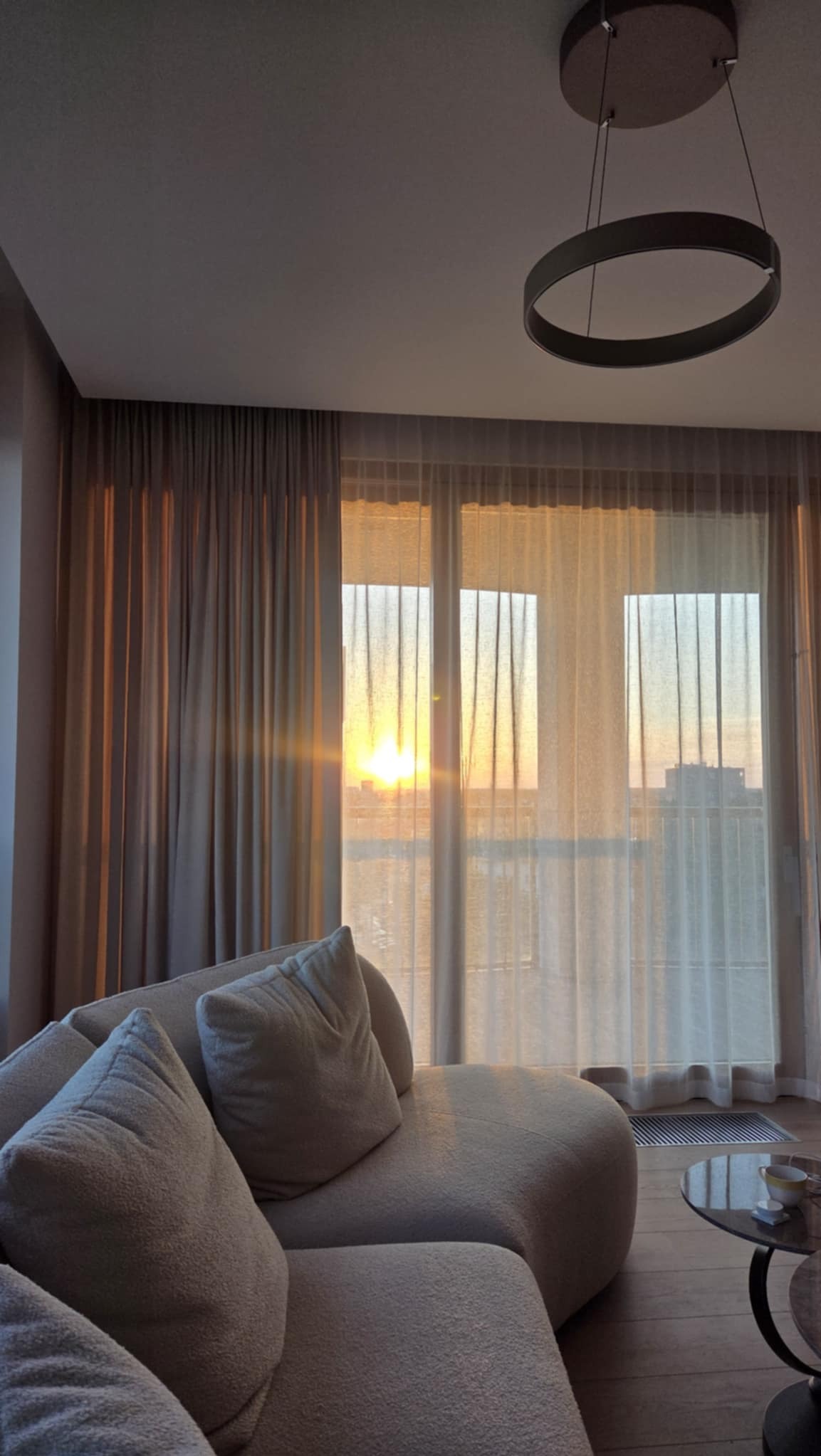 Beżowe, długie zasłony w salonie z widokiem na zachód słońca, przez okno. Wnętrze dopełnia nowoczesna lampa i sofa z poduszkami.