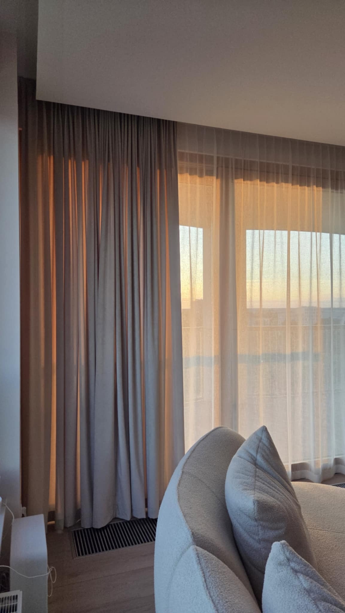 Beżowe, długie zasłony i firany w oknie z widokiem na zachód słońca. U dołu widoczny fragment sofy z dekoracyjnymi poduszkami. Przytulne wnętrze.