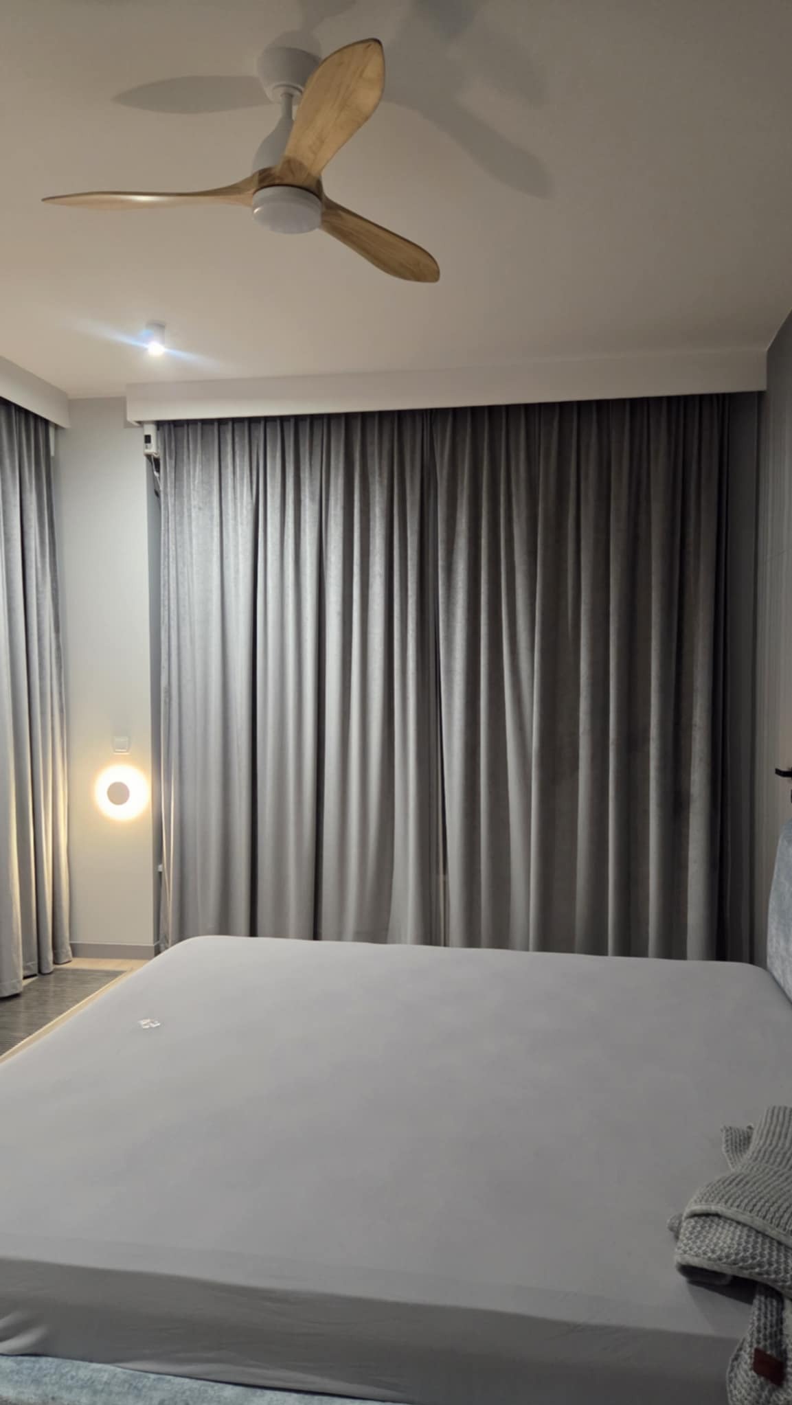 Elegancka sypialnia z szarymi, welurowymi zasłonami, drewnianym wentylatorem sufitowym i minimalistycznym oświetleniem ściennym. Subtelna kolorystyka i nowoczesny design.