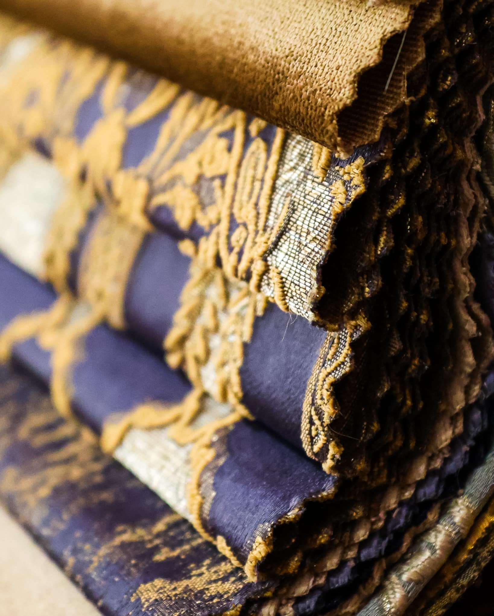 Stos próbek tkanin zasłonowych w odcieniach fioletu i złota, prezentujący bogate wzornictwo i faktury materiałów, idealny do wyboru zasłon na zamówienie.