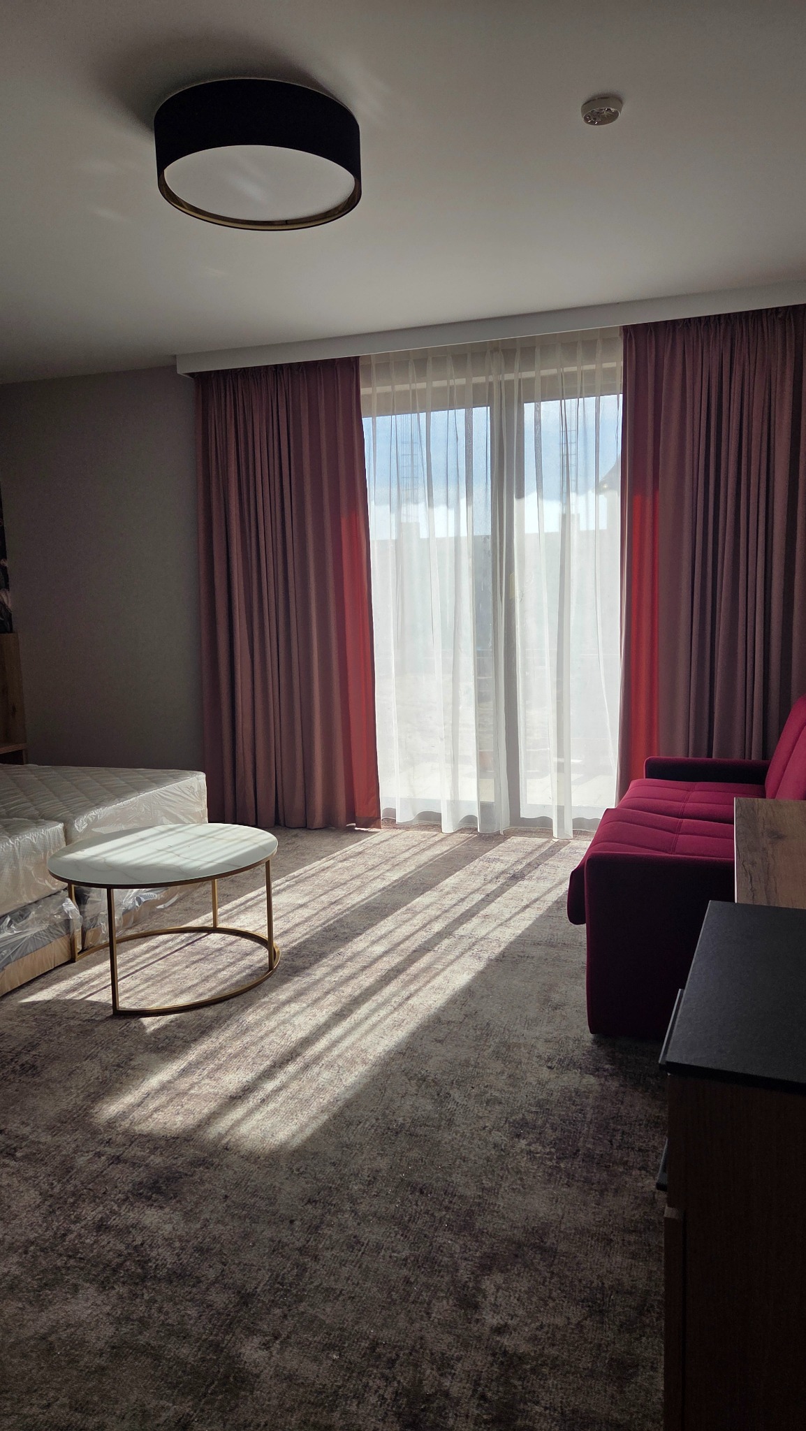 Nowoczesny salon z zasłonami w odcieniach różu i bieli, wpadające promienie słoneczne, okrągły stolik kawowy, sofa. Aranżacja okienna z efektem miękkiego światła.