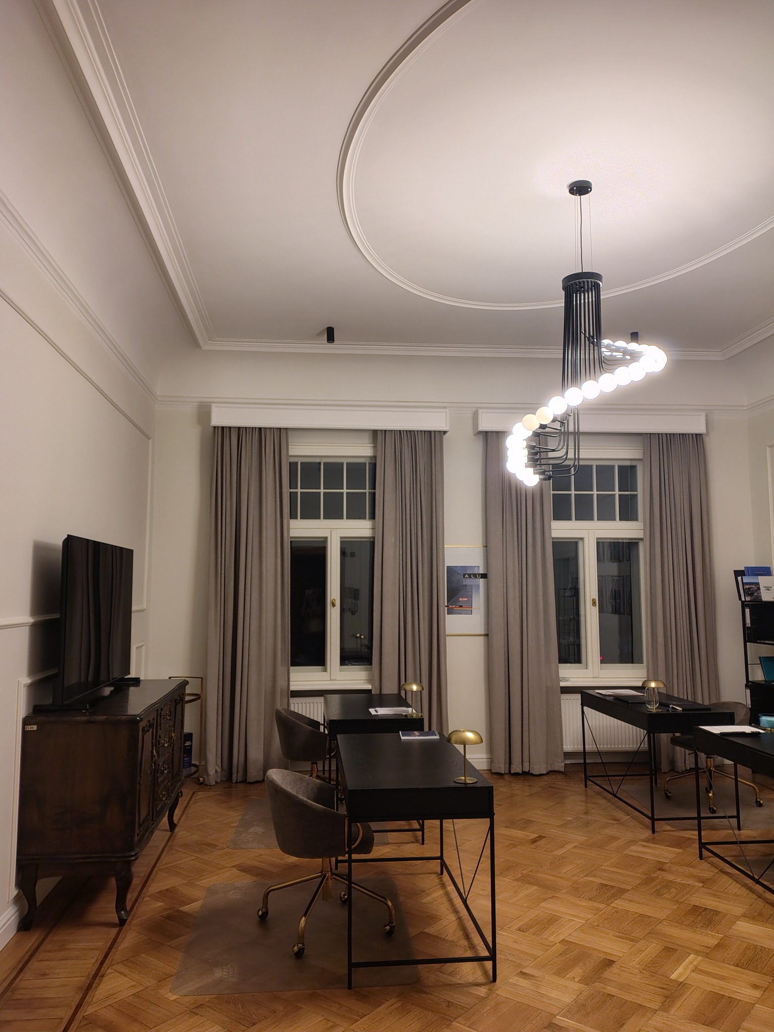 Eleganckie, szare zasłony w klasycznym wnętrzu z parkietem, biurkami i designerską lampą. Długie, proste zasłony w oknach w biurze.