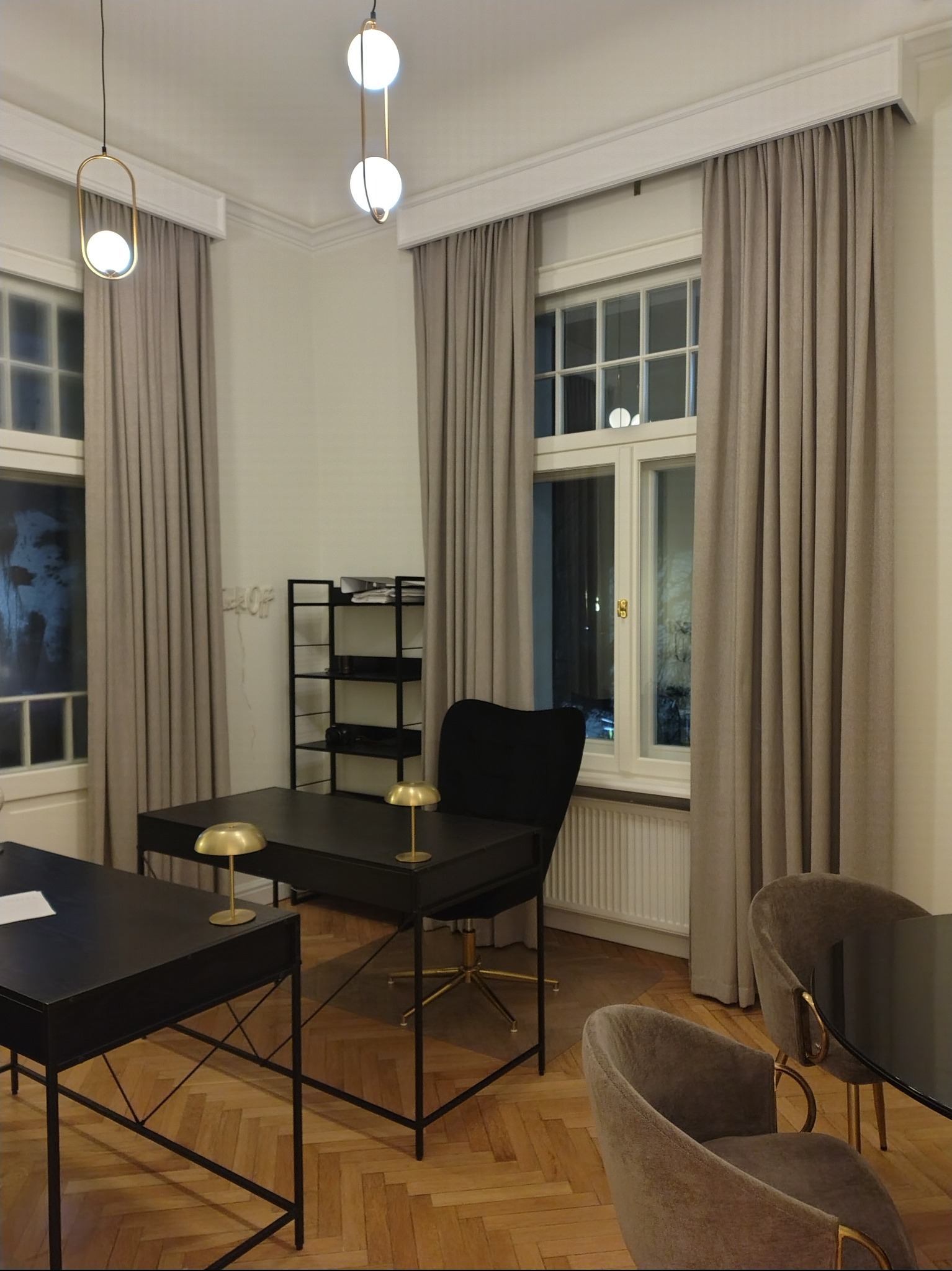 Eleganckie, beżowe zasłony w gabinecie z czarnymi biurkami i złotymi lampkami, widoczne przez okno nocne miasto. Klasyczny wystrój wnętrza.