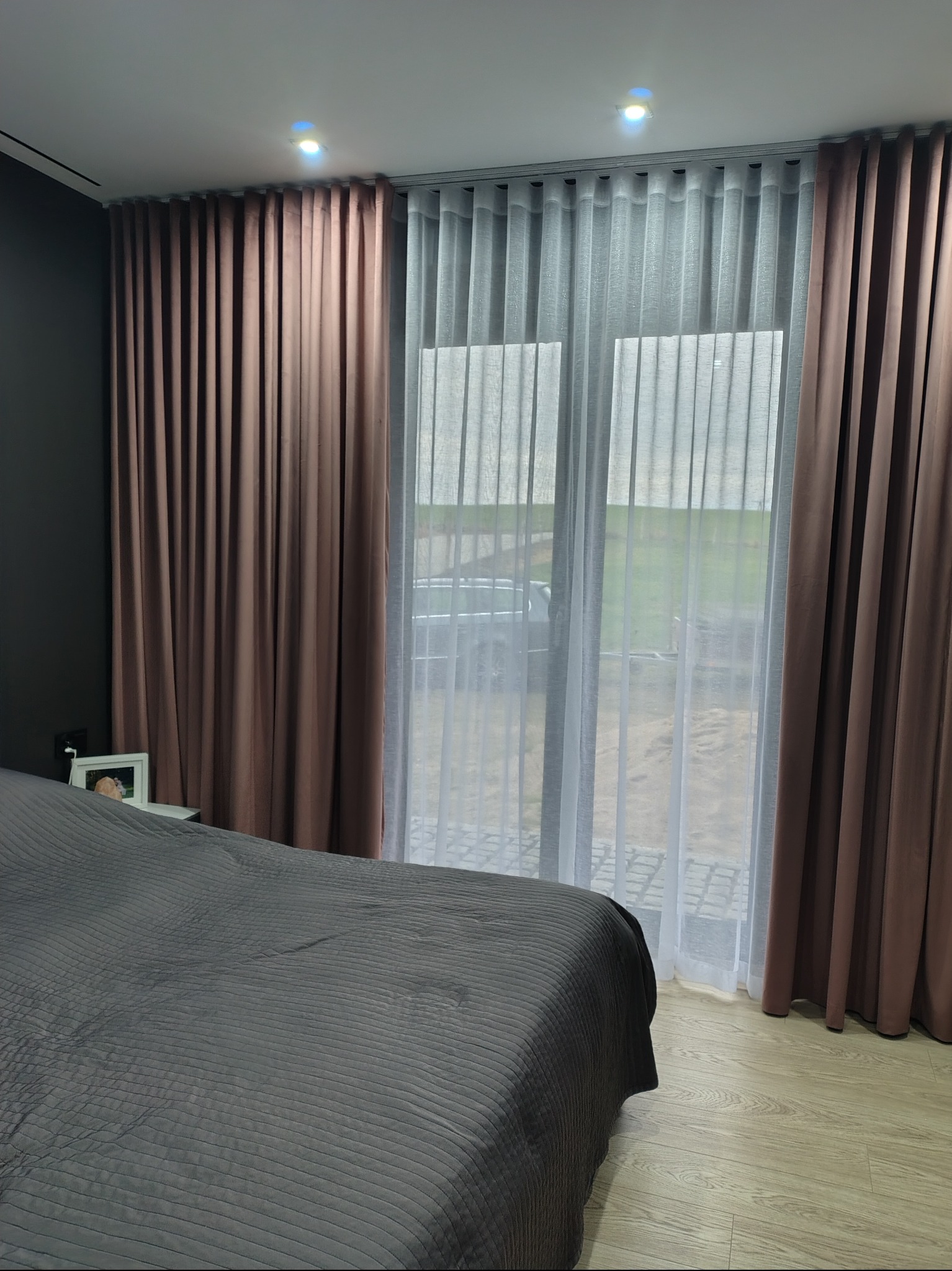 Eleganckie, brązowe zasłony zaciemniające i białe, zwiewne firany w sypialni z szarym łóżkiem. Widok na zewnątrz przez okno.