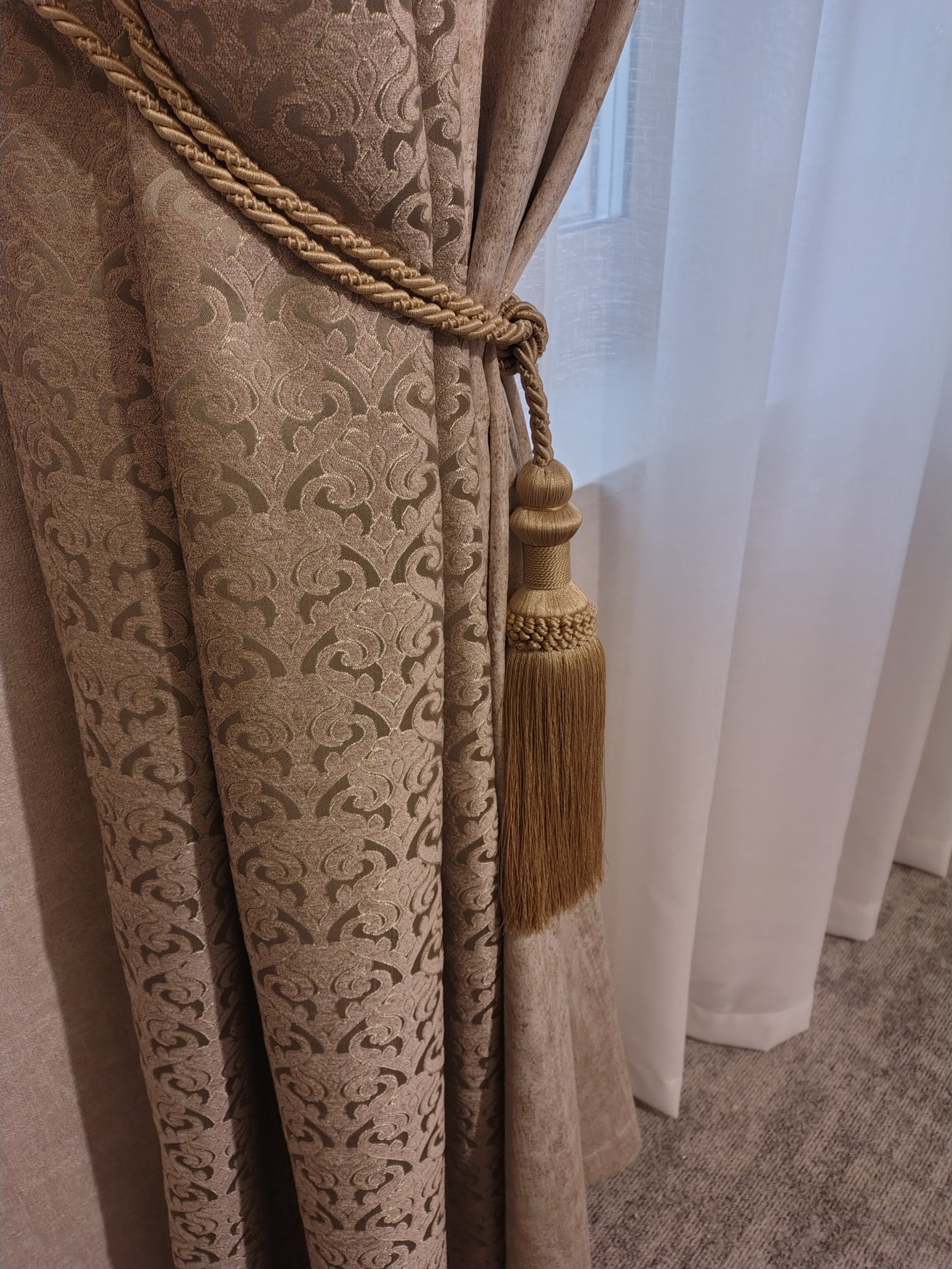 Elegancka, brązowa zasłona z dekoracyjnym wzorem, upięta złotym sznurem z ozdobnym chwostem. W tle widoczne białe, delikatne firany i fragment okna.