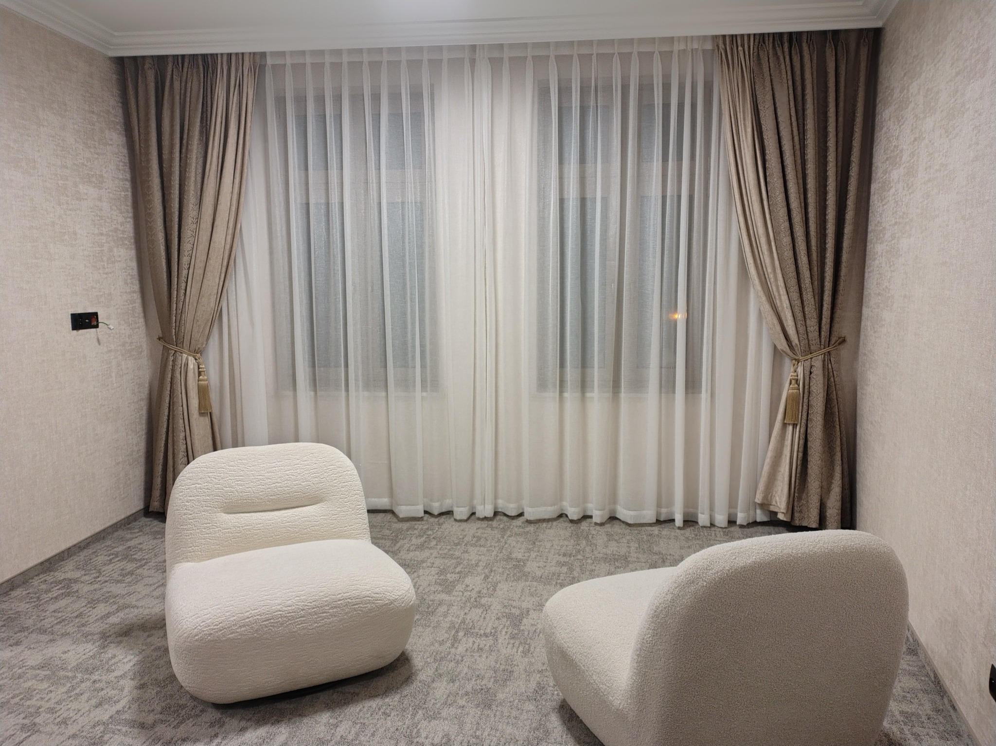 Eleganckie, beżowe zasłony z delikatnym wzorem, upięte sznurami, w zestawieniu z białymi, zwiewnymi firanami. Dwa białe fotele boucle na tle szarej podłogi.