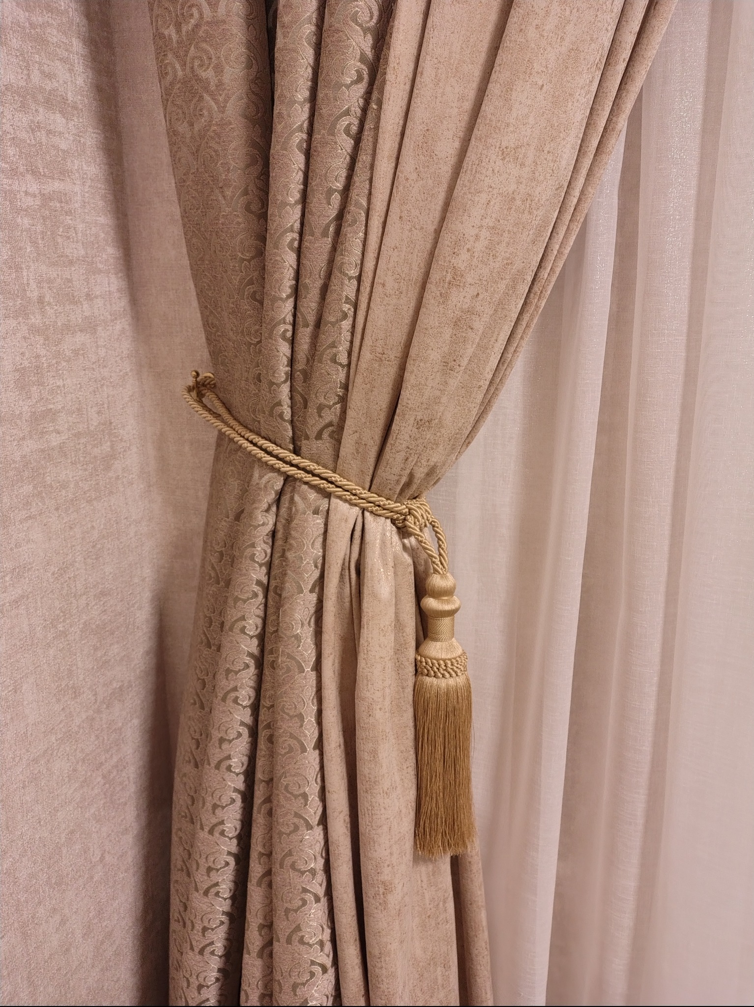 Beżowa, wzorzysta zasłona upięta złotym sznurem z ozdobnym chwostem. W tle delikatna, jasna firana i ściana w podobnym odcieniu. Dekoracja okienna w klasycznym stylu.