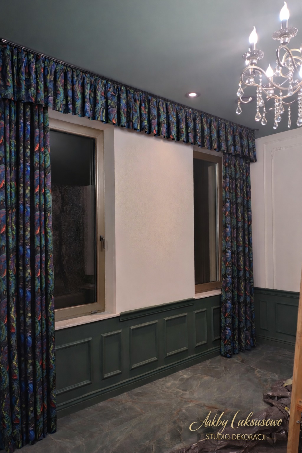 Eleganckie, długie zasłony z wzorzystego materiału, dopasowane do karnisza i wystroju wnętrza z ciemnozieloną boazerią i marmurową podłogą. Widoczny żyrandol.
