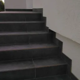 Zewnętrzne schody wyłożone ciemnymi, matowymi płytkami, z widocznym fragmentem elewacji budynku w tle.