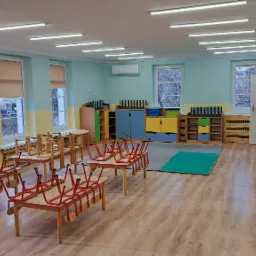 Sala przedszkolna z drewnianą podłogą, pomalowana na jasne kolory, z oknami z roletami, na stołach odwrócone krzesła, w tle kolorowe szafki i dywanik.